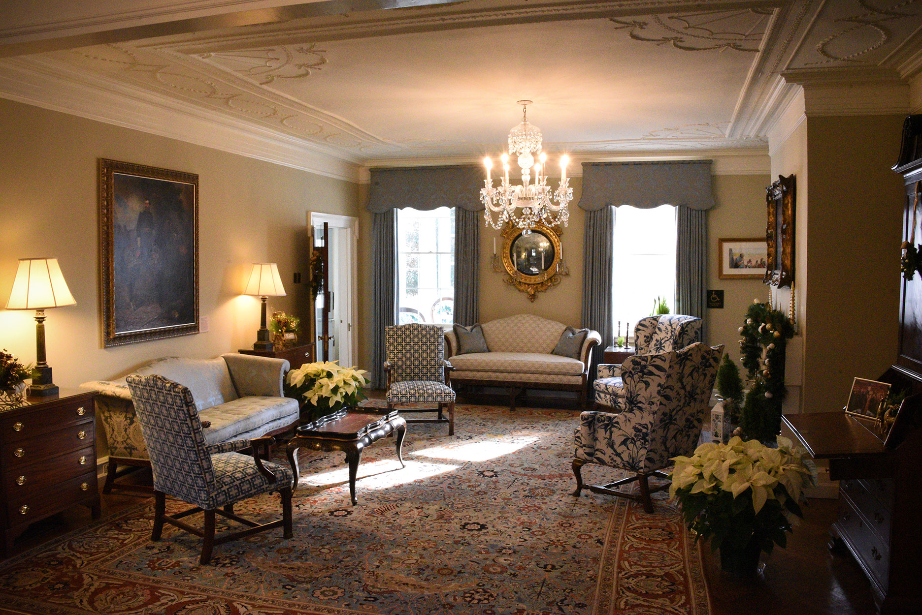 Sunlight pours through Parlor windows where Warren Garden Club graces the room with white poinsettias, gold and green seasonal decor. Elegant details, furniture and carpet fill the room.