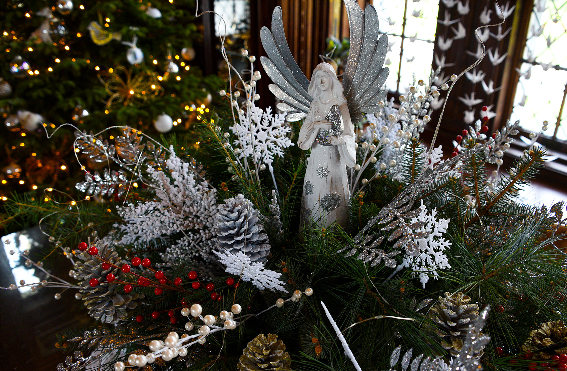 A display on the desk in the Library, an angel atop a spray of greenery, conifers and berries. A Christmas tree in the background.