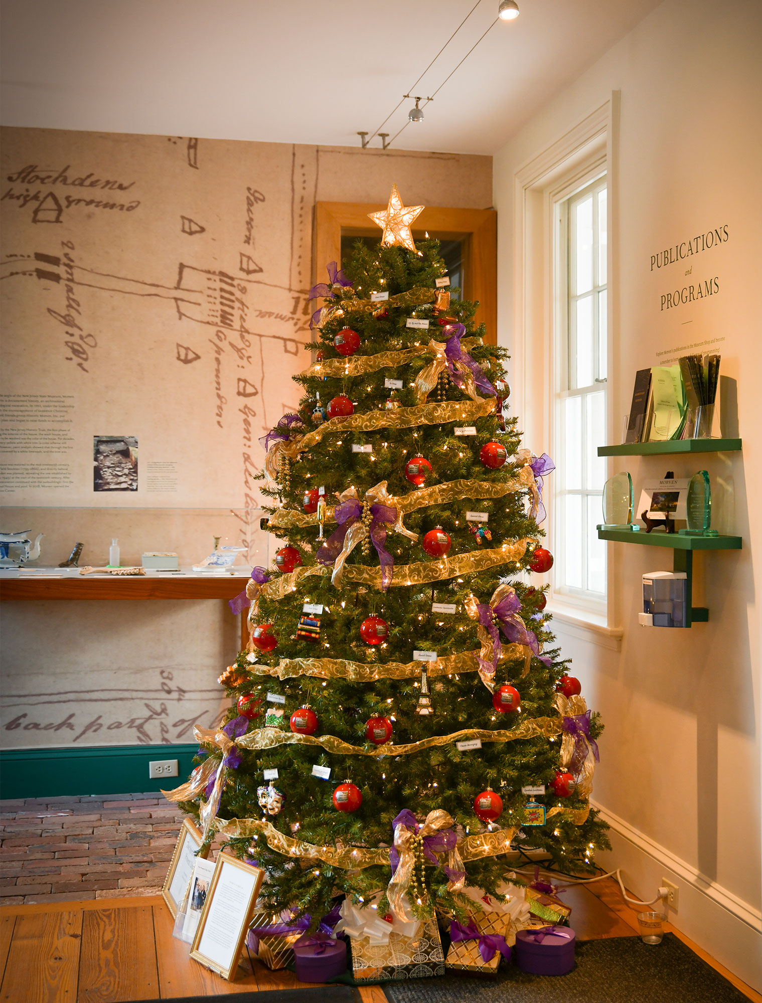 The “Spirit of the Present Day” Christmas tree stands at the rear entryway to Morven. A festive gold ribbon wraps around many colors of special and cheerful ornaments and bows. Museum displays can be seen  to the right and to the left.
