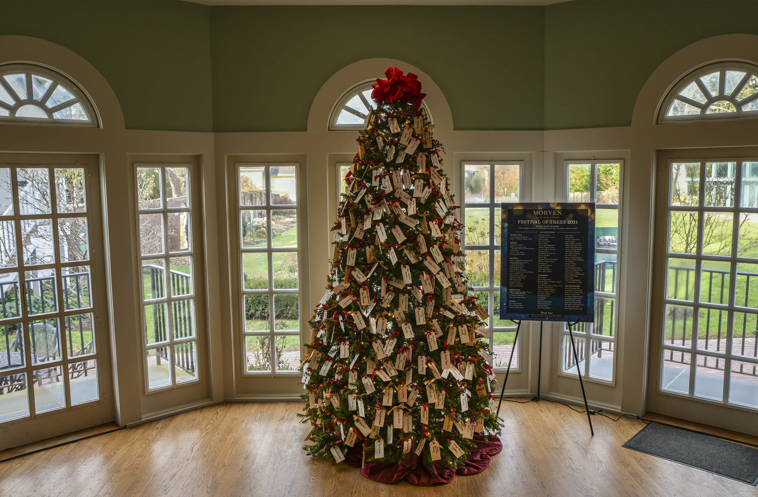 A Christmas tree adorned with dozens of tags with peoples names stands in the middle of a green room. The rear garden area can be seen through windows and doors surrounding the display.