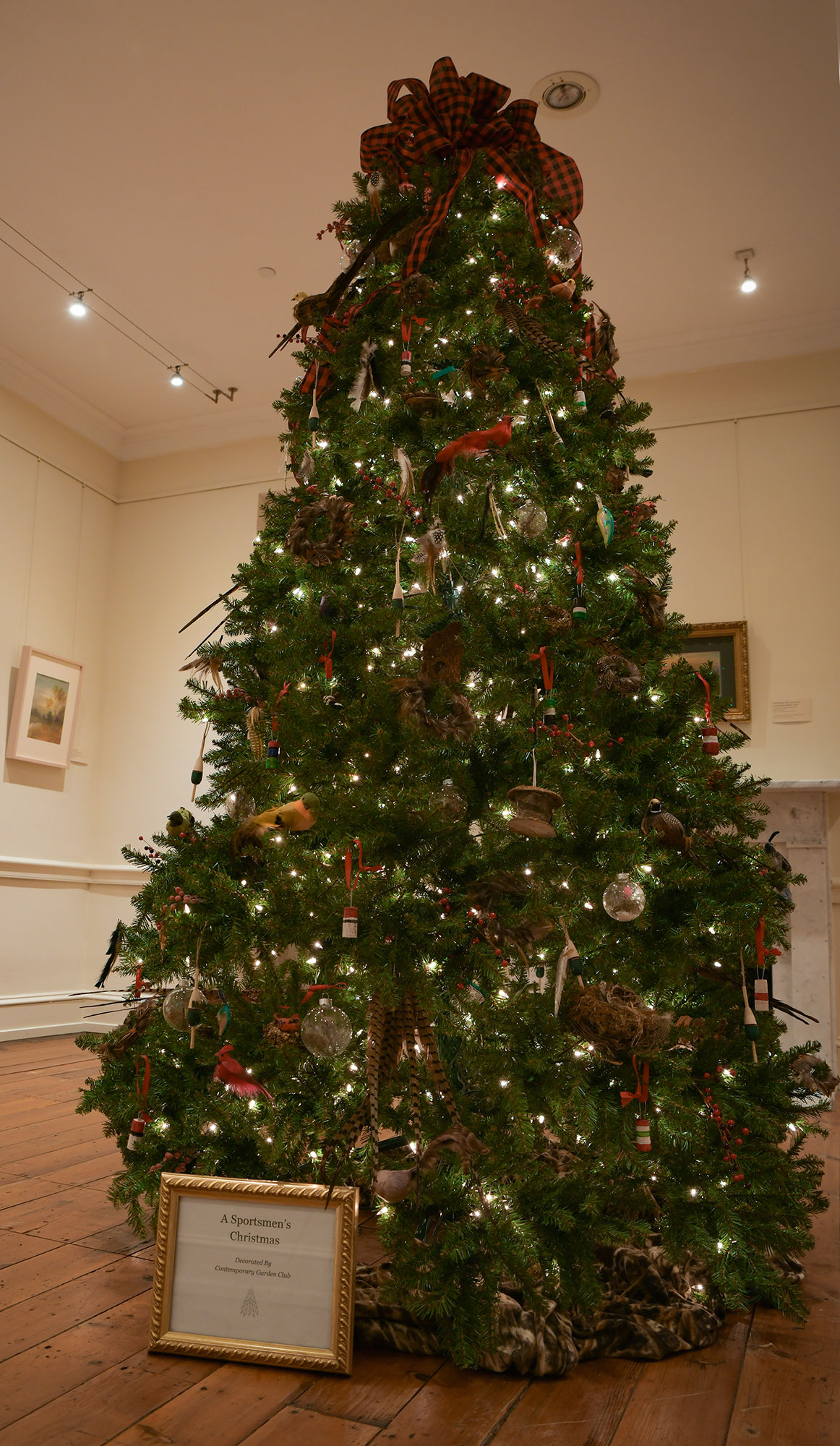 A Christmas tree, in a beige room with a wooden floor and paintings in the background, is decorated with feathers, bird ornaments and a red and black plaid bow at the top. A framed display at the front reads Sportsmens Christmas by the Contemporary 